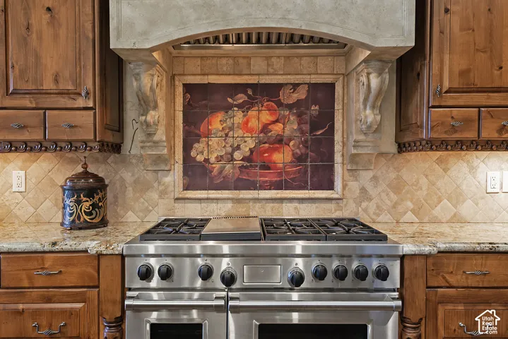 Stainless steel range with grill, decorative tile backsplash, granite countertops, custom wood cabinetry, and a stone-clad range hood with corbels