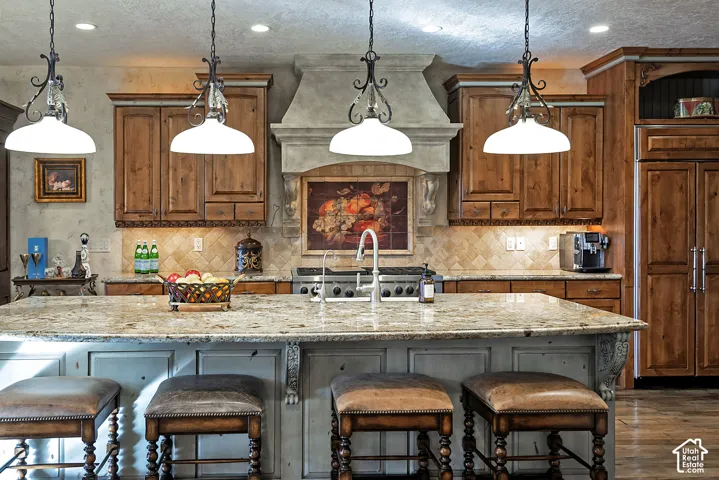Kitchen featuring a stone-clad range hood, wood cabinetry, tile backsplash, and a large island with a stone countertop