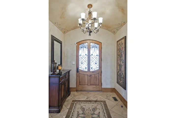 Arched wooden entry door with decorative iron and glass inserts, tiled flooring with an inlaid pattern, vaulted ceiling with a decorative light fixture, and textured wall finishes