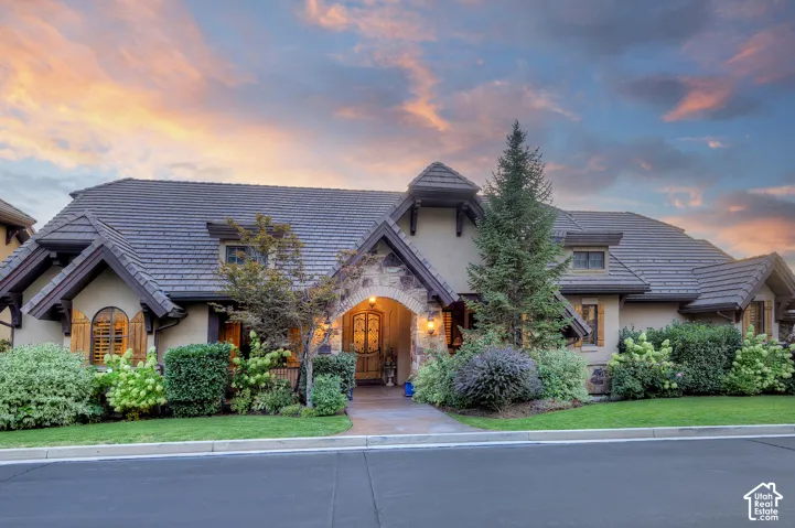 Stucco exterior with stone accents, arched entry, dark tiled roof, dormer windows, and established landscaping