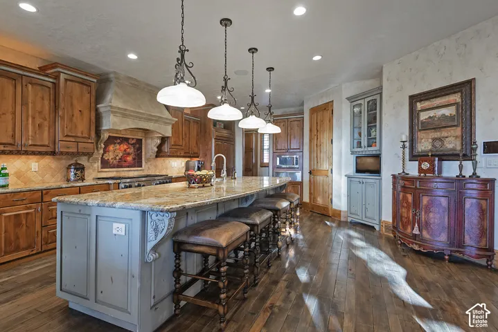 Gourmet kitchen featuring a large central island with seating, granite countertops, custom wood cabinetry, a tiled backsplash, and recessed and pendant lighting