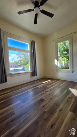 Spacious room featuring wood-finish flooring, multiple windows, a ceiling fan with integrated lighting, and neutral wall tones