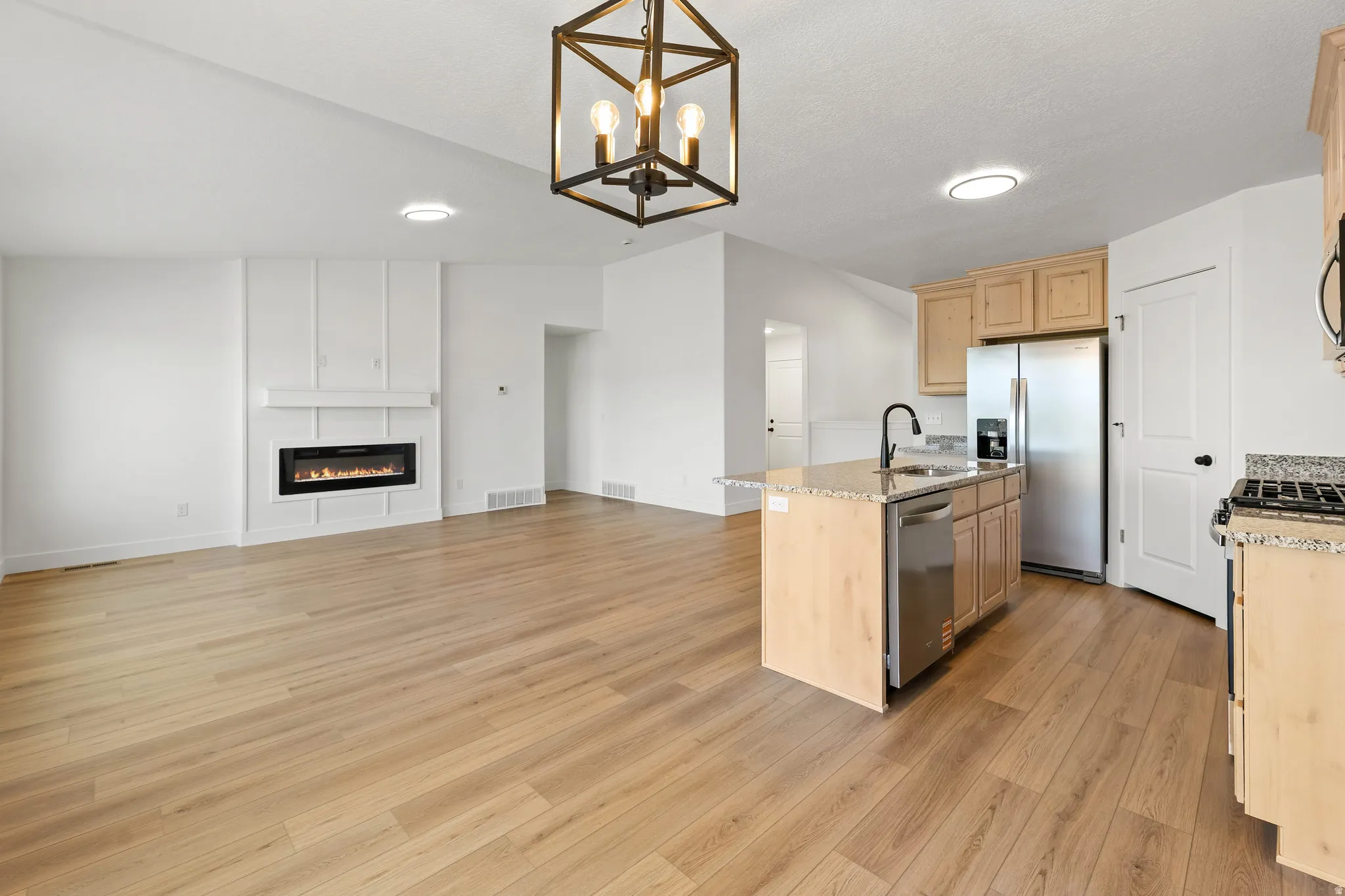Open-concept living area featuring wood-finish flooring, a contemporary fireplace with a decorative surround, a kitchen island with stone countertops, stainless steel appliances, and a vaulted ceiling with a modern light fixture