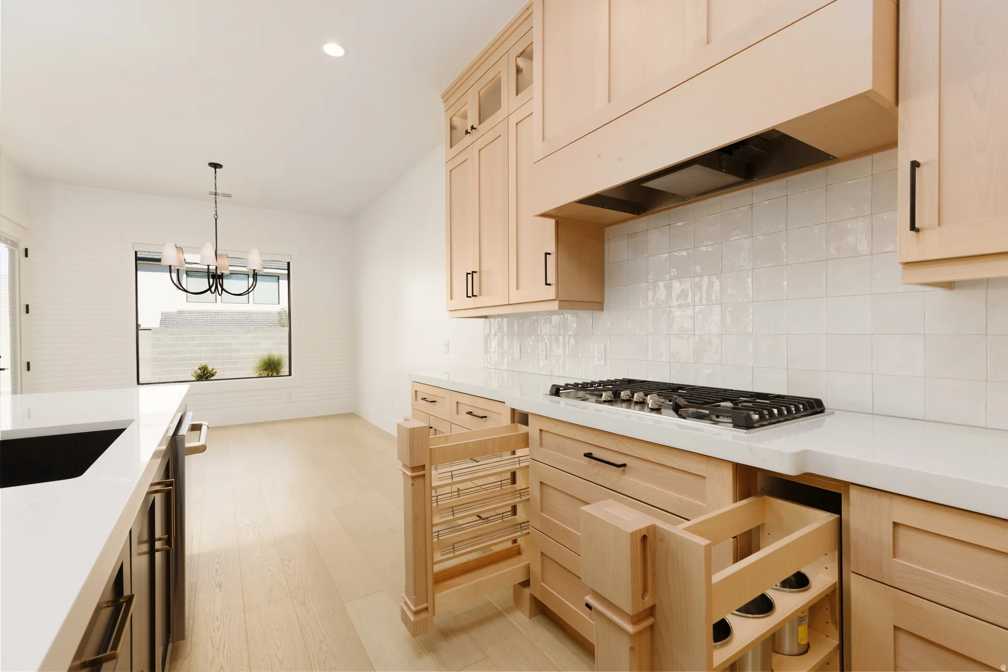Light wood-finish cabinetry, white countertops, built-in cooktop, textured tile backsplash, and wood-finish flooring