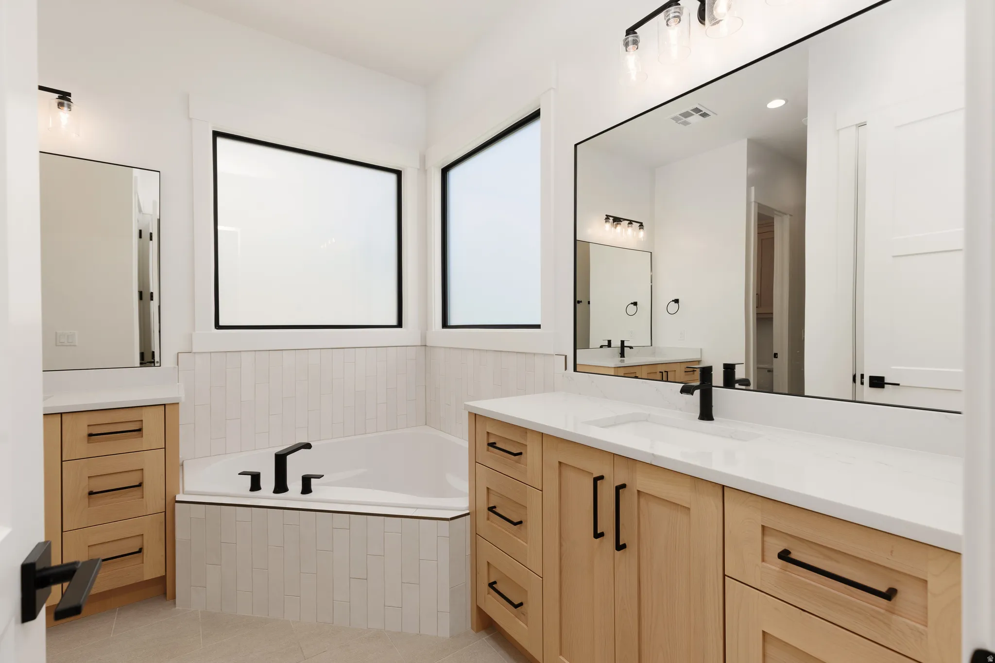 Soaking tub with vertical tile surround, wood-finish cabinetry, white countertops, and matte black fixtures