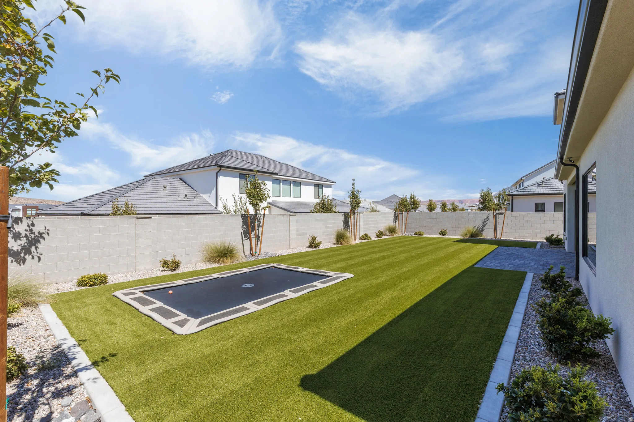 Expansive backyard featuring a manicured artificial turf lawn, stone paver patio, integrated trampoline, and a block perimeter fence