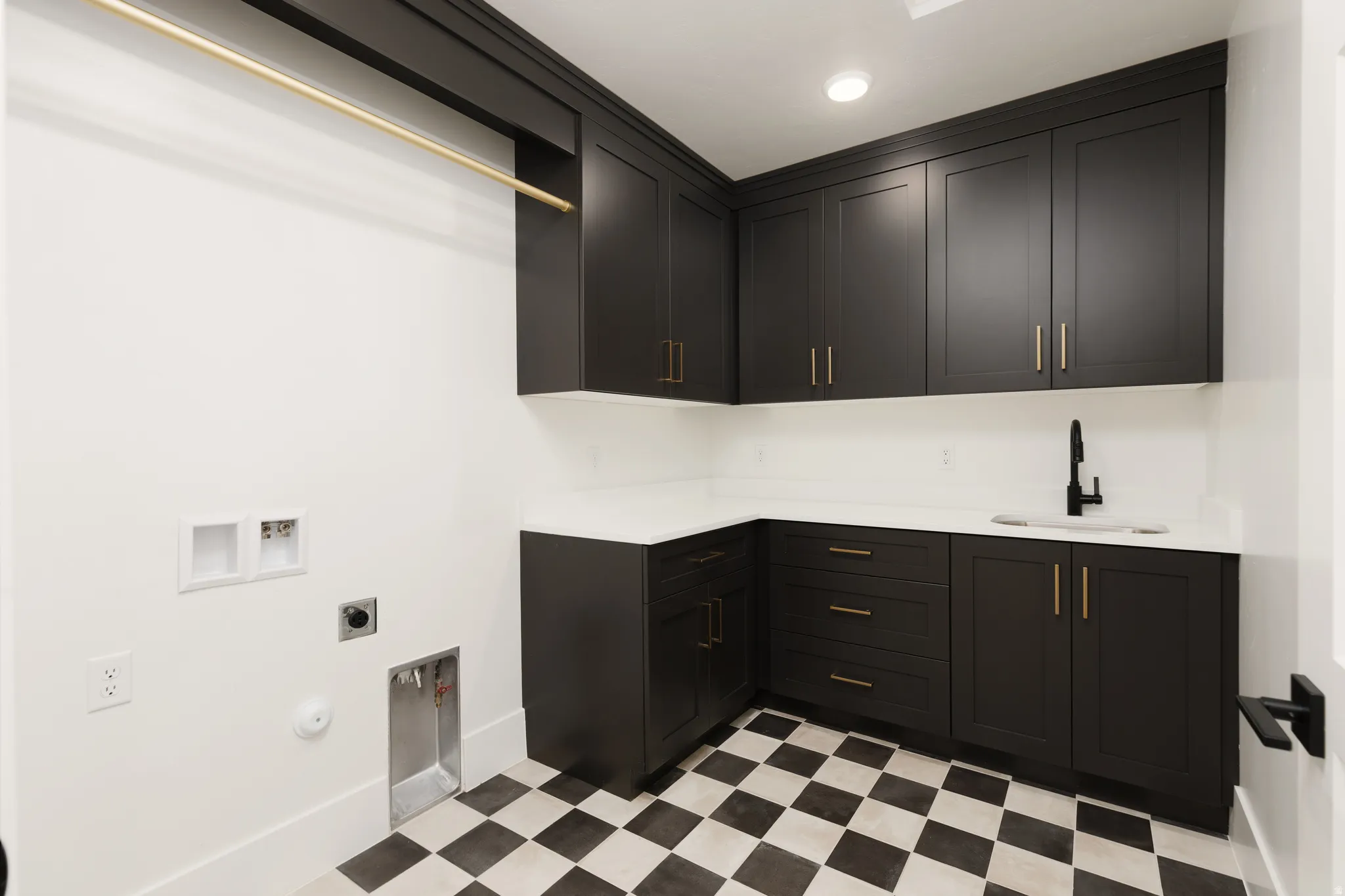 Black and white checkered flooring, dark cabinetry with gold-tone hardware, white countertops, wall-mounted drying rod, and a single-basin sink with a matte black faucet