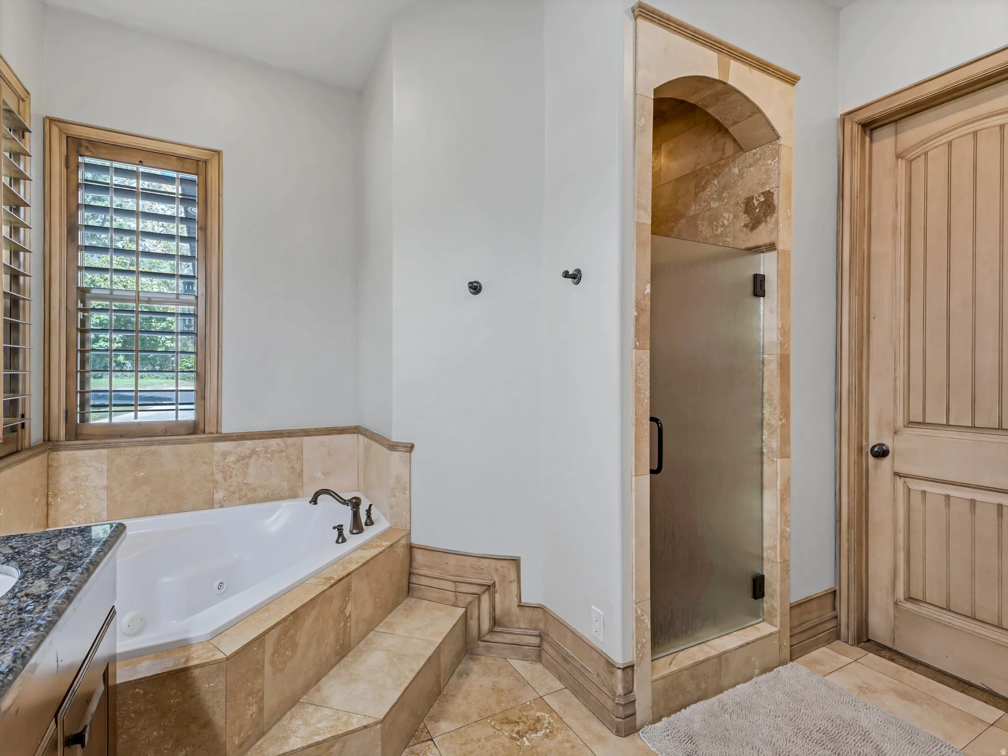 Jetted soaking tub, walk-in shower with frosted glass door, natural stone tile surround, wood-frame window with plantation shutters, and wood-paneled door