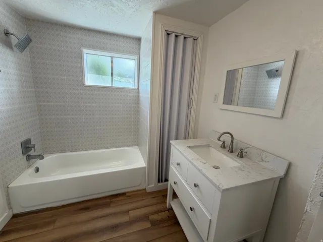 White vanity with a stone-finish countertop, wood-finish flooring, tiled shower surround, and a white bathtub