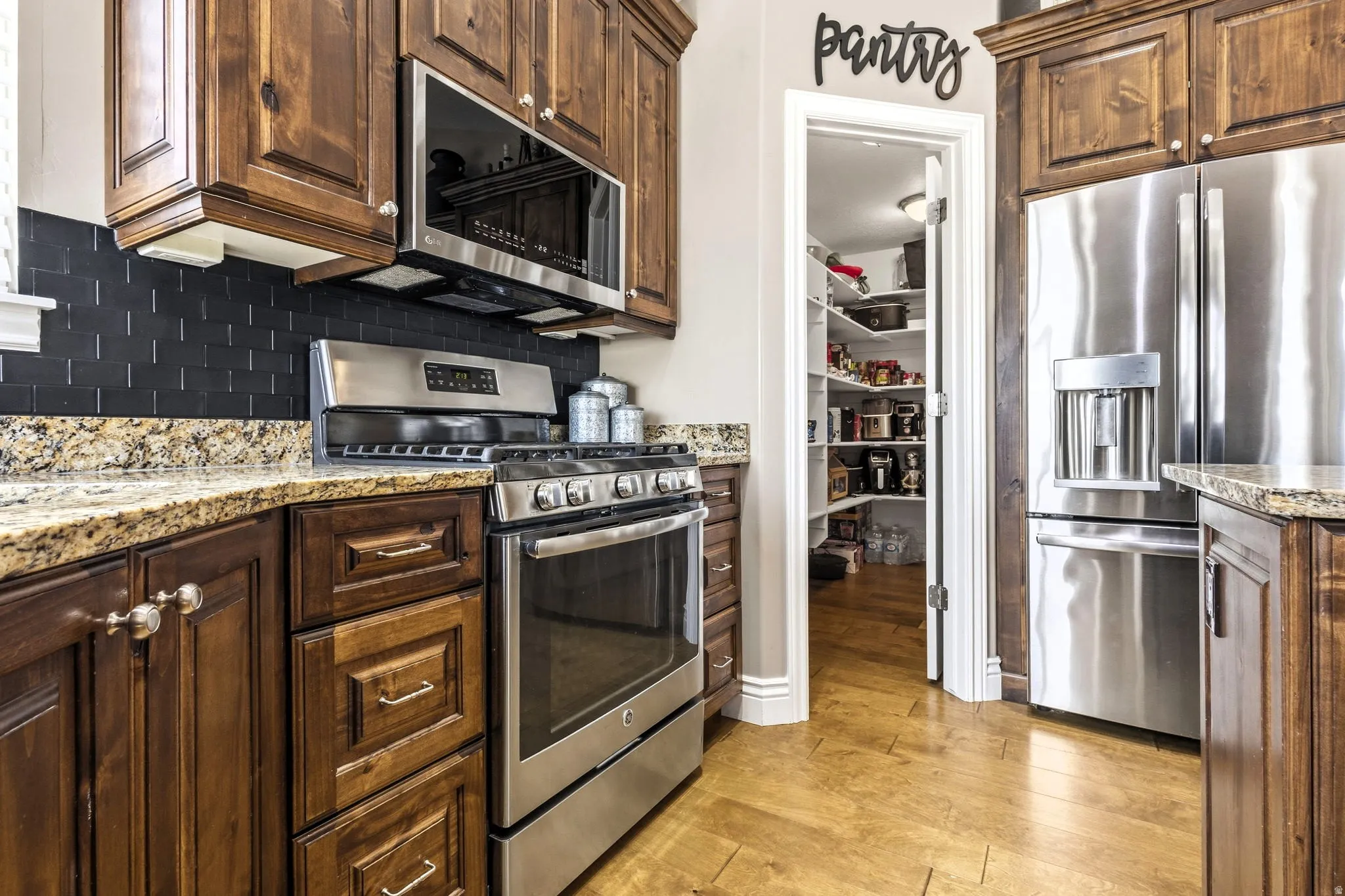Wood-finish cabinetry, stainless steel appliances, granite countertops, dark tile backsplash, and a walk-in pantry