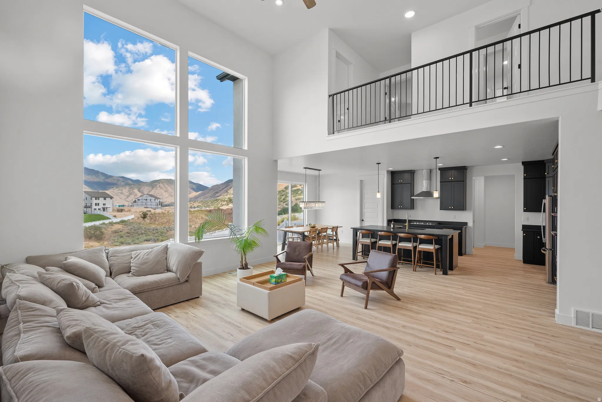 Expansive living area with soaring ceilings, large picture windows, wood-finish flooring, a kitchen island with seating, and a lofted space with iron railings
