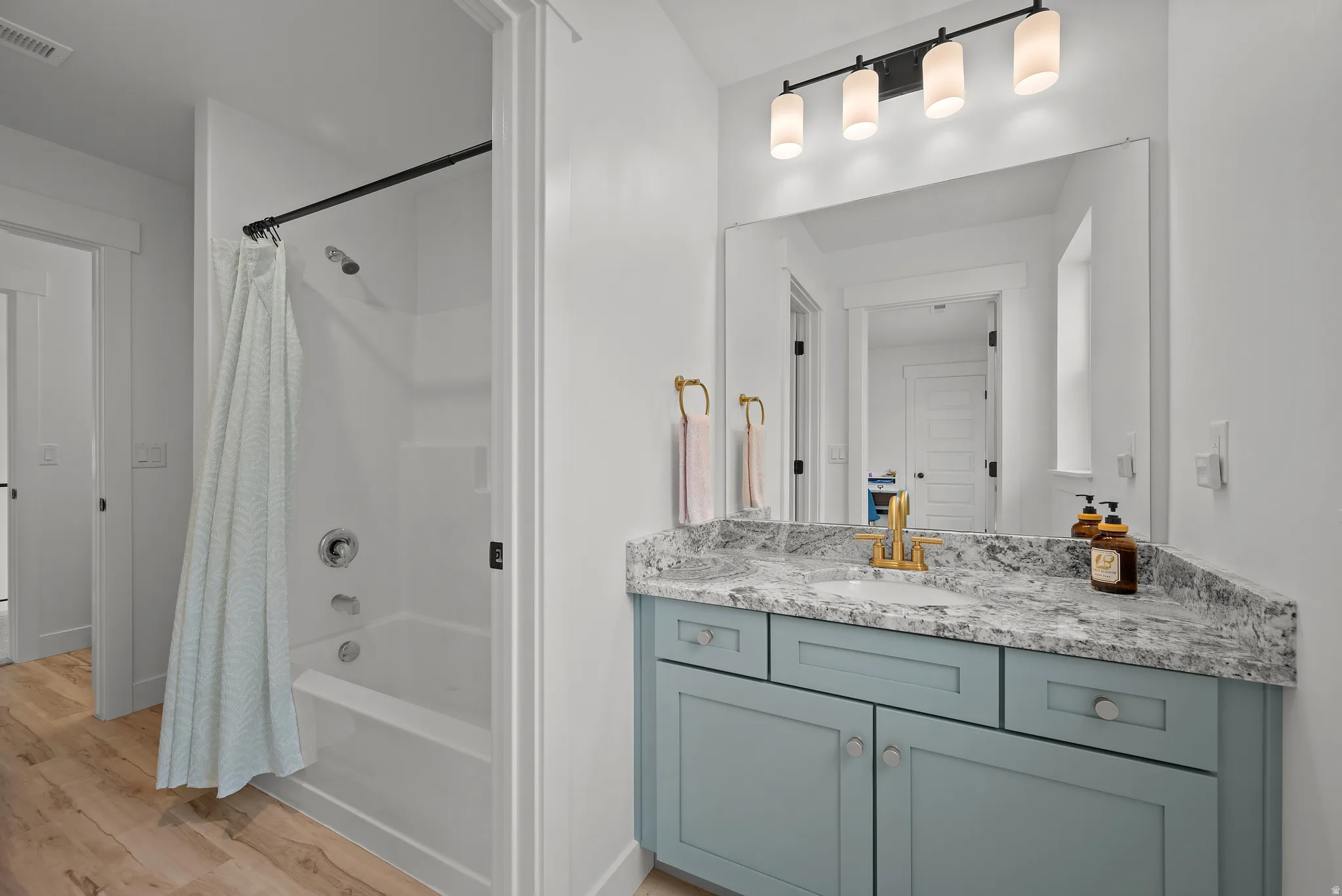 White bathtub and shower combination with chrome fixtures, wood-finish flooring, a vanity with a light blue cabinet, a granite-style countertop, and a rectangular mirror with an overhead light fixture