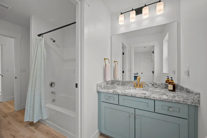 White bathtub and shower combination with chrome fixtures, wood-finish flooring, a vanity with a light blue cabinet, a granite-style countertop, and a rectangular mirror with an overhead light fixture
