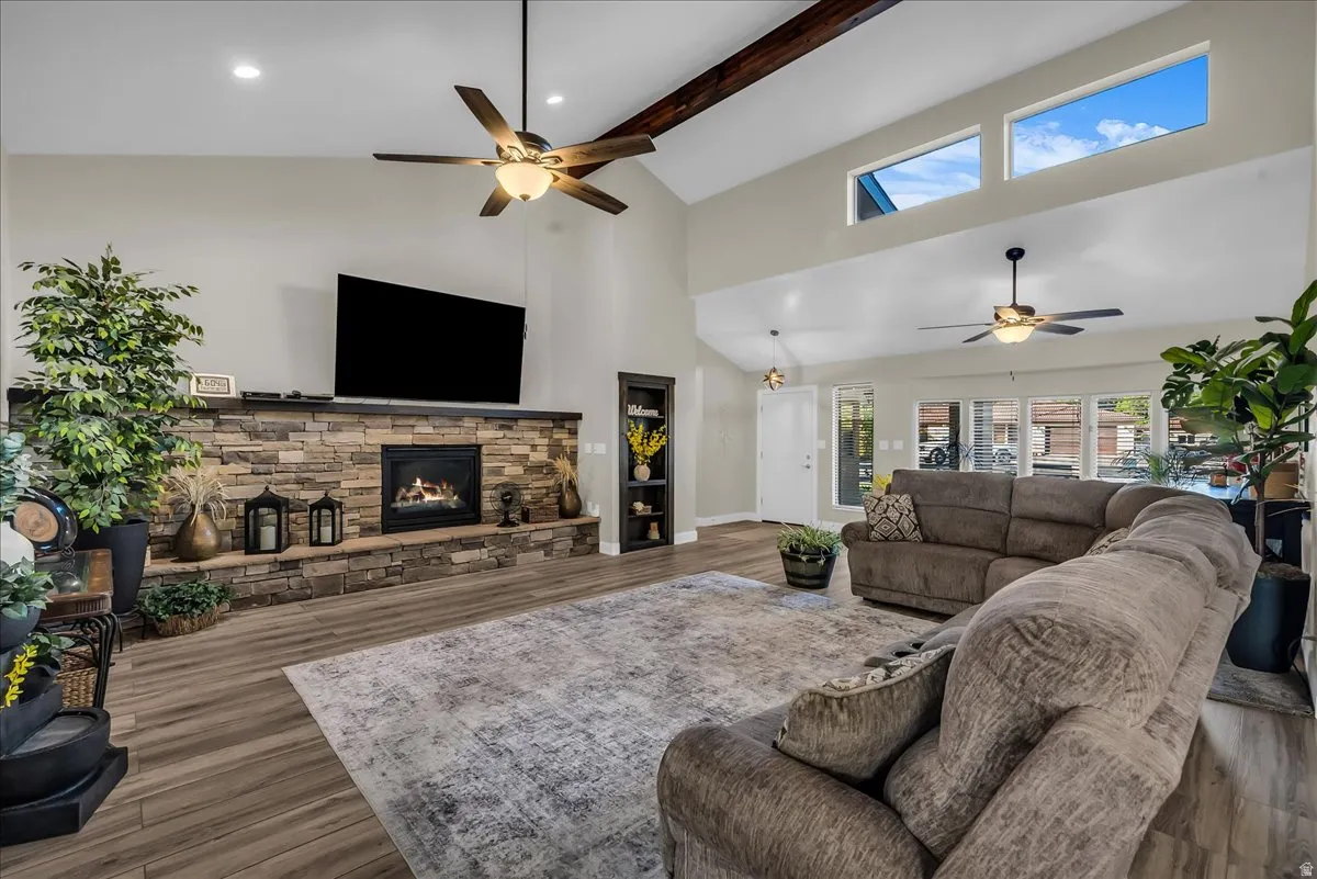 Vaulted ceiling with exposed wood beam, stacked stone fireplace with hearth, wood-finish flooring, built-in shelving, and transom windows
