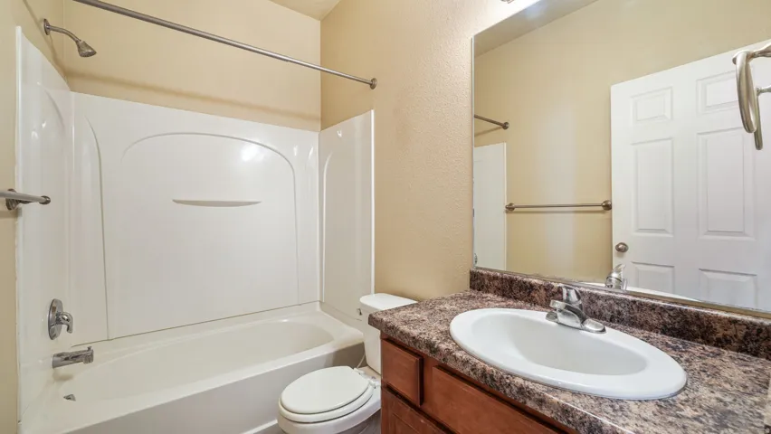 White bath and shower combination with chrome fixtures, wood-finish vanity with an integrated sink, patterned countertop, and an overhead vanity light