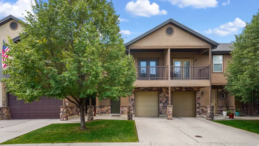 Stucco exterior with stone accents, two-car garage, upper-level balcony, and mature trees