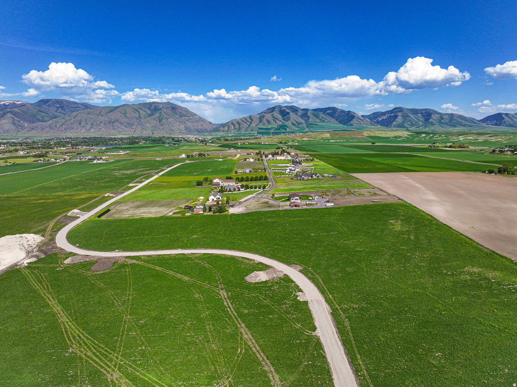 Expansive green fields, mountain backdrop, residential cluster, winding paved road, additional development area