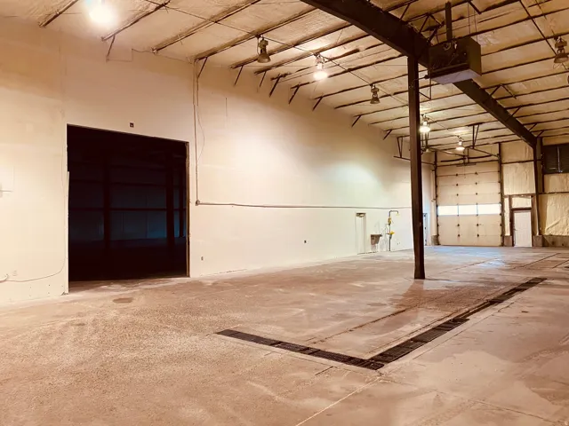 High-bay warehouse space featuring exposed steel beams, concrete flooring, multiple overhead lighting fixtures, floor drain and 16' roll-up door