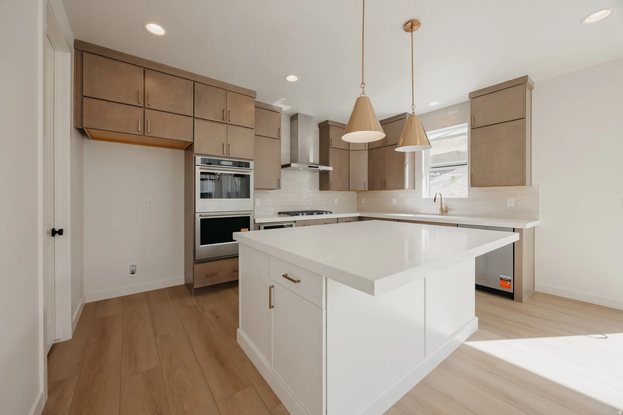 Kitchen featuring a center island with a light-toned countertop, stainless steel double oven, stainless steel range hood, light wood-finish flooring, and pendant lighting