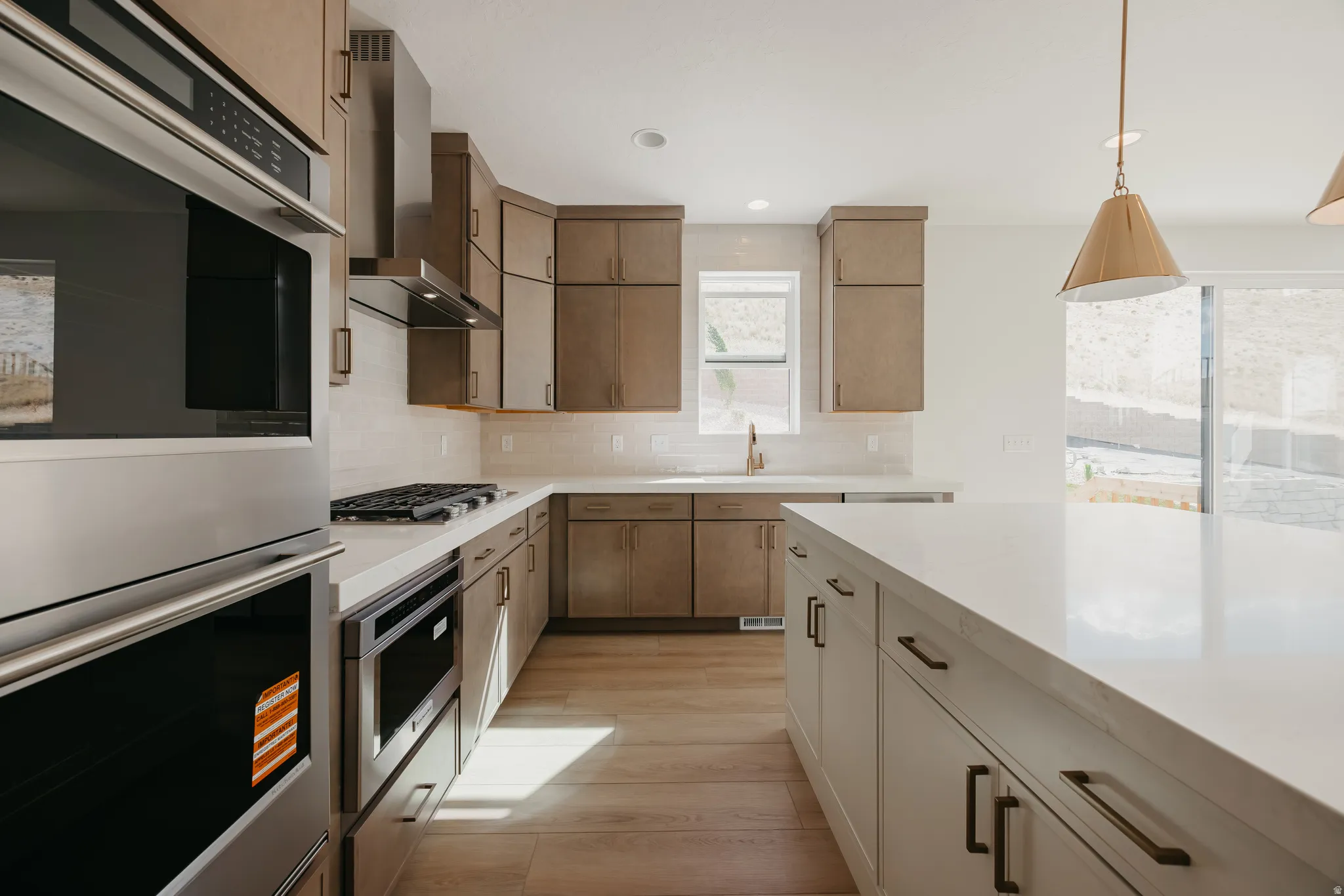 Kitchen with wood-finish flooring, built-in double ovens, stainless steel range hood, white countertops, and a kitchen island with storage