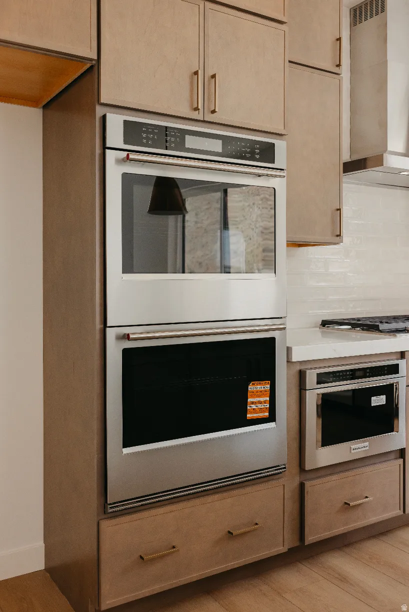 Integrated stainless steel double ovens, custom cabinetry with brushed gold hardware, wood-finish flooring, white subway tile backsplash, and recessed stainless steel microwave