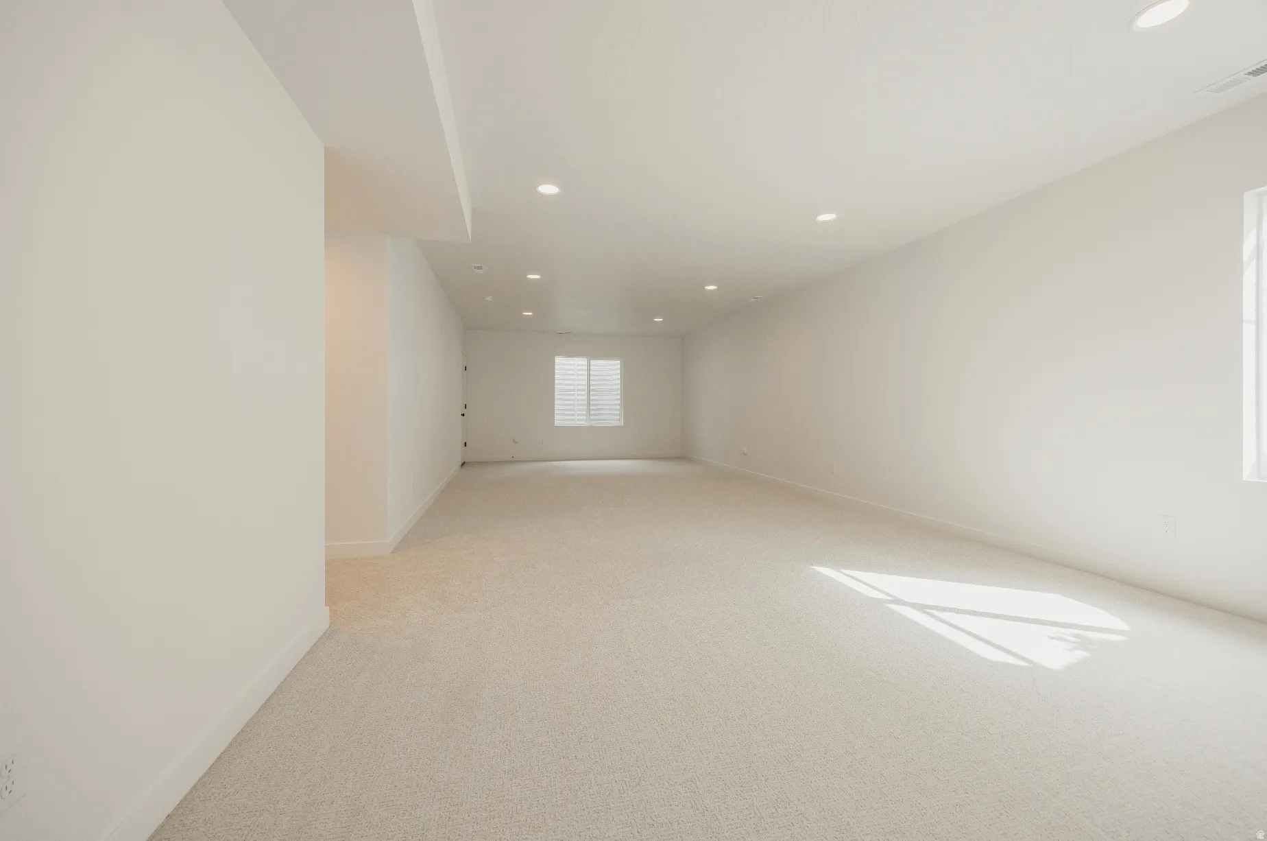 Recessed lighting illuminates a spacious room featuring light-toned carpeting and a window with horizontal blinds