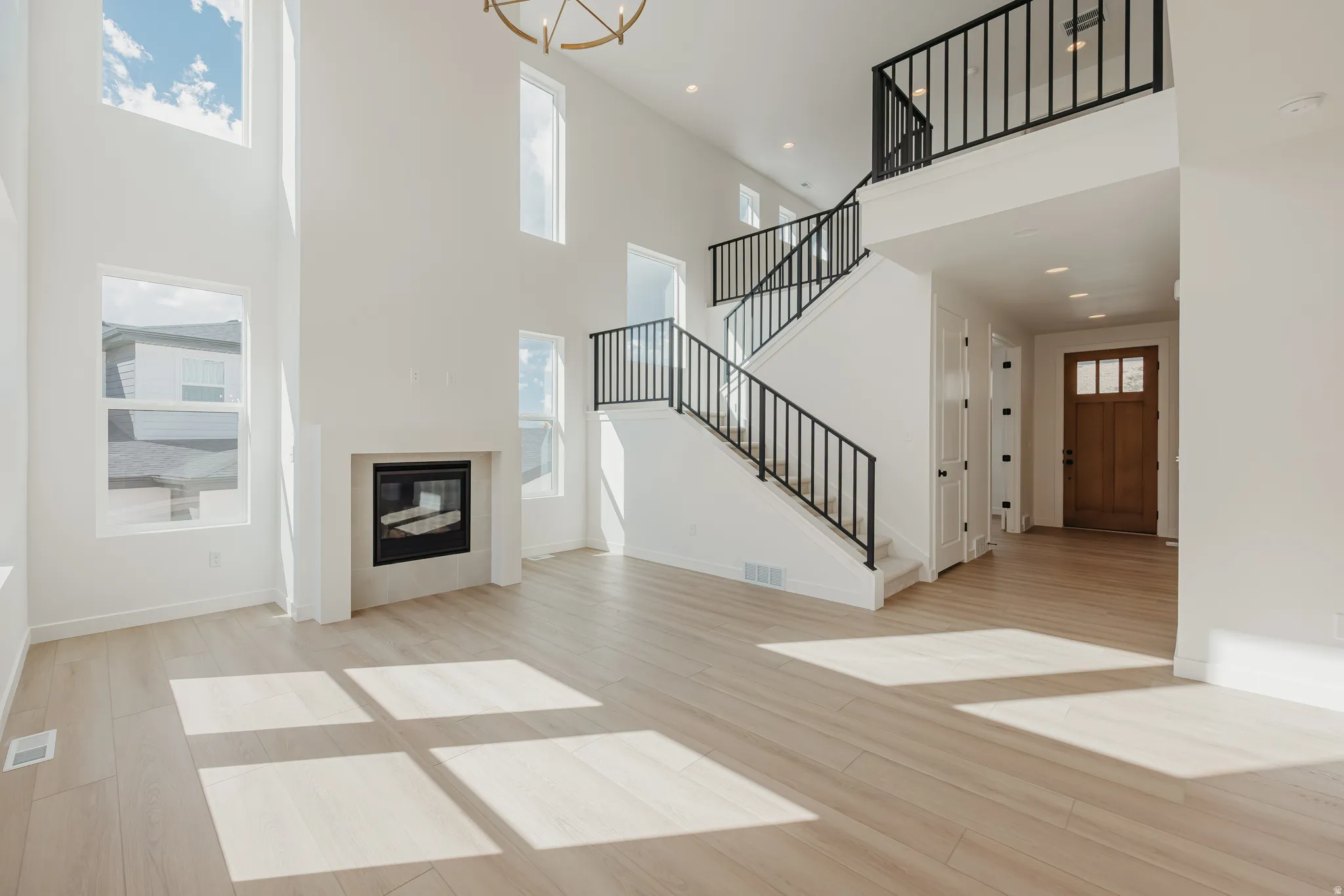 Two-story living area featuring a fireplace, wood-finish flooring, a grand staircase with metal railings, a statement chandelier, and an entryway with a wood door