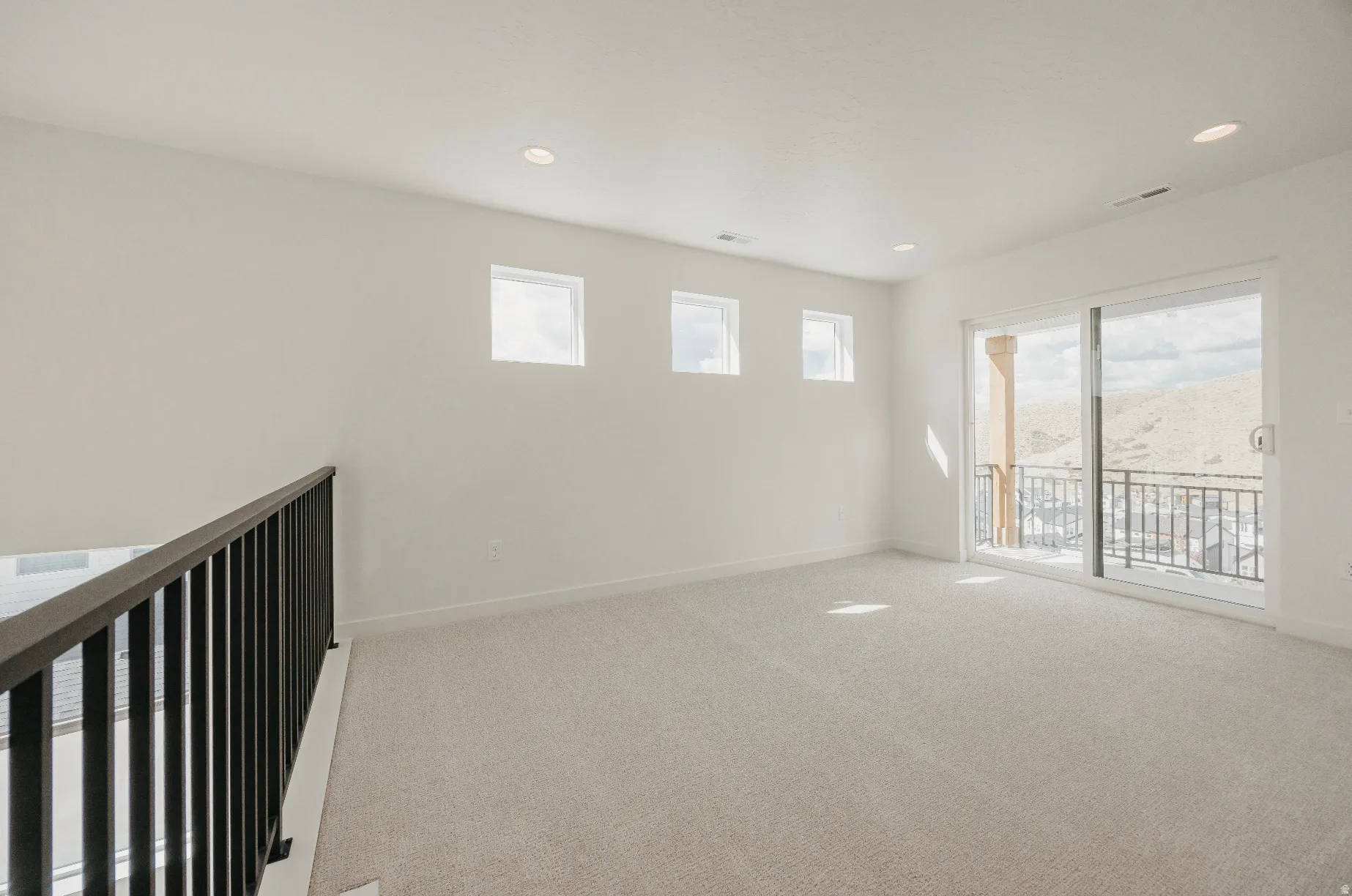 Spacious room featuring light-colored carpeting, recessed lighting, multiple transom windows, and a sliding glass door leading to a balcony with metal railings