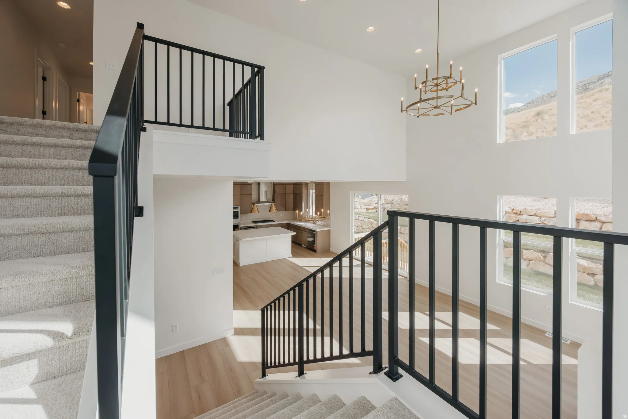 Two-story ceiling, multiple windows, carpeted staircase, black metal railings, and a prominent chandelier