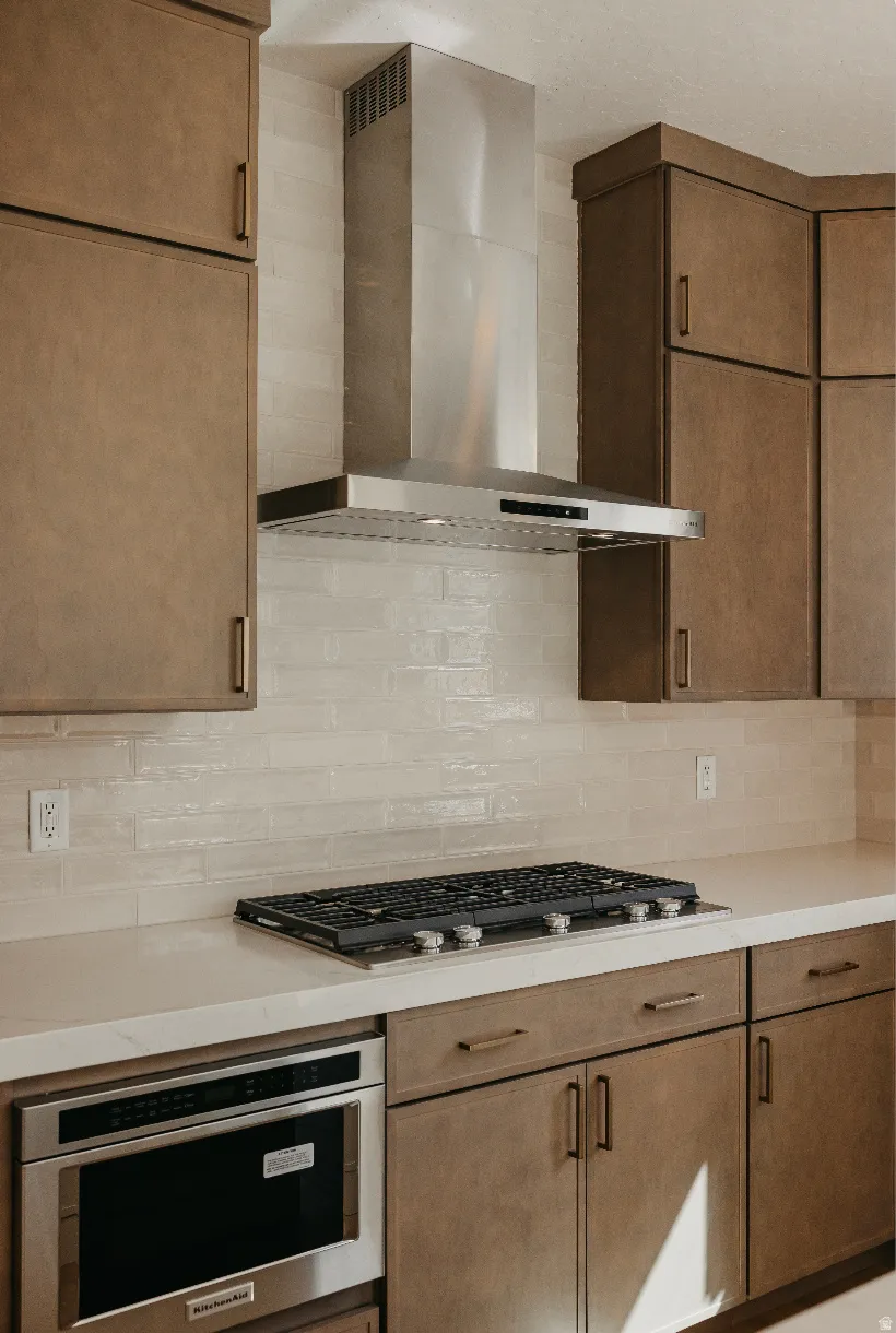 Stainless steel range hood, built-in gas cooktop, integrated microwave, white subway tile backsplash, and light-toned cabinetry