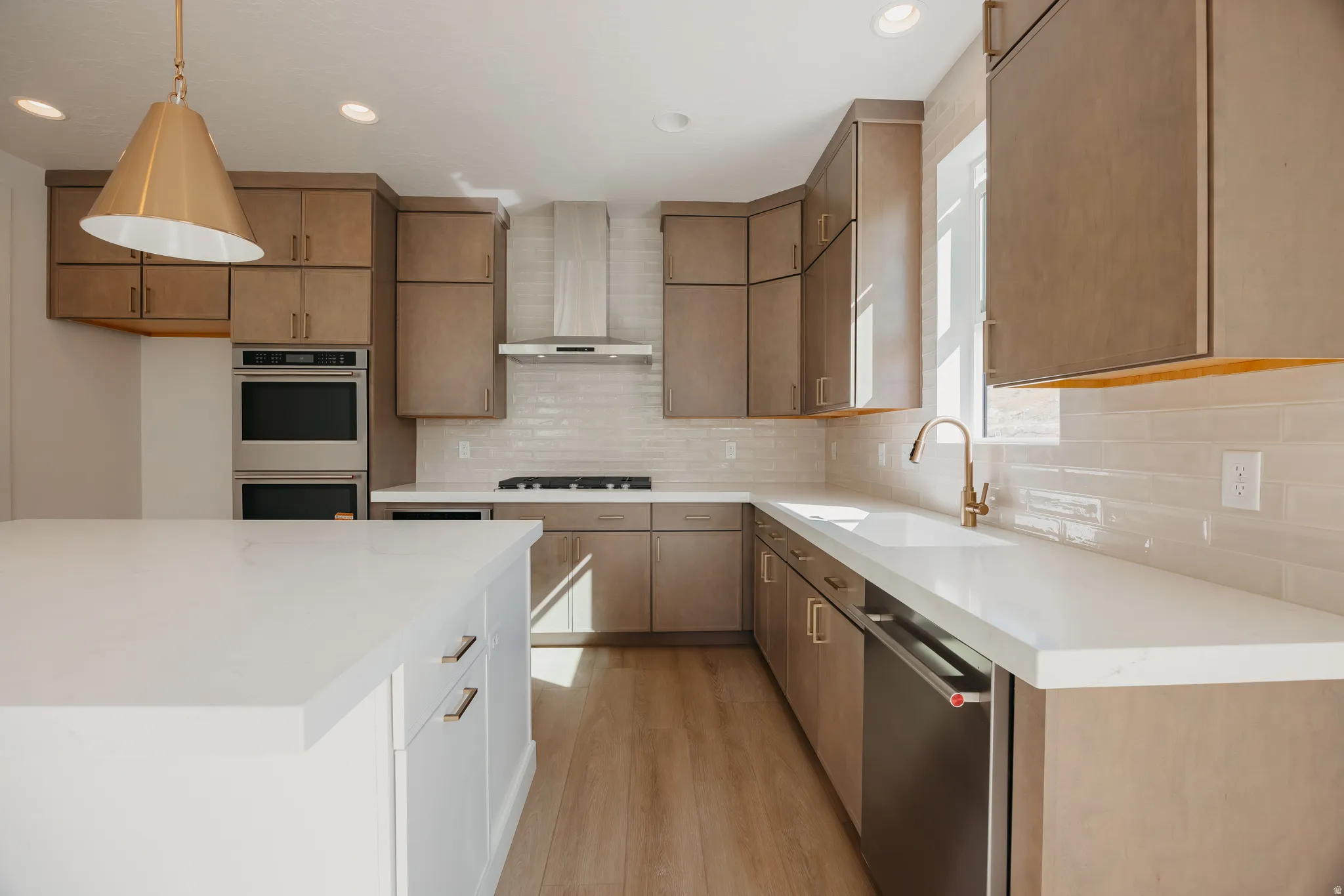 Modern kitchen featuring wood-finish cabinetry, white quartz countertops, stainless steel appliances including dual wall ovens and gas cooktop, subway tile backsplash, and warm wood-finish flooring throughout