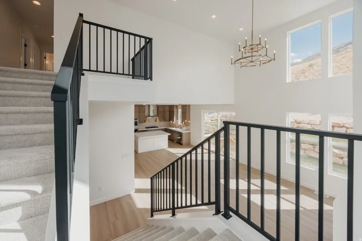 Two-story ceiling, multiple windows, carpeted staircase, black metal railings, and a prominent chandelier