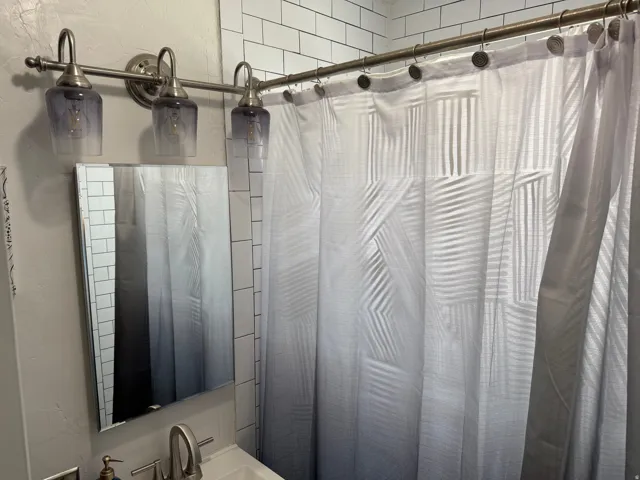 Updated vanity lighting fixture with three glass shades, rectangular framed mirror, and subway tile shower surround
