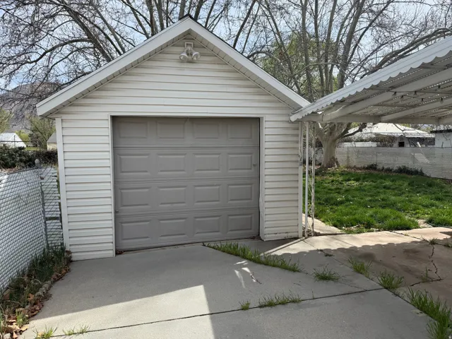 Detached garage with vinyl siding and a single garage door, featuring an exterior light fixture, concrete driveway, and a chain-link fence