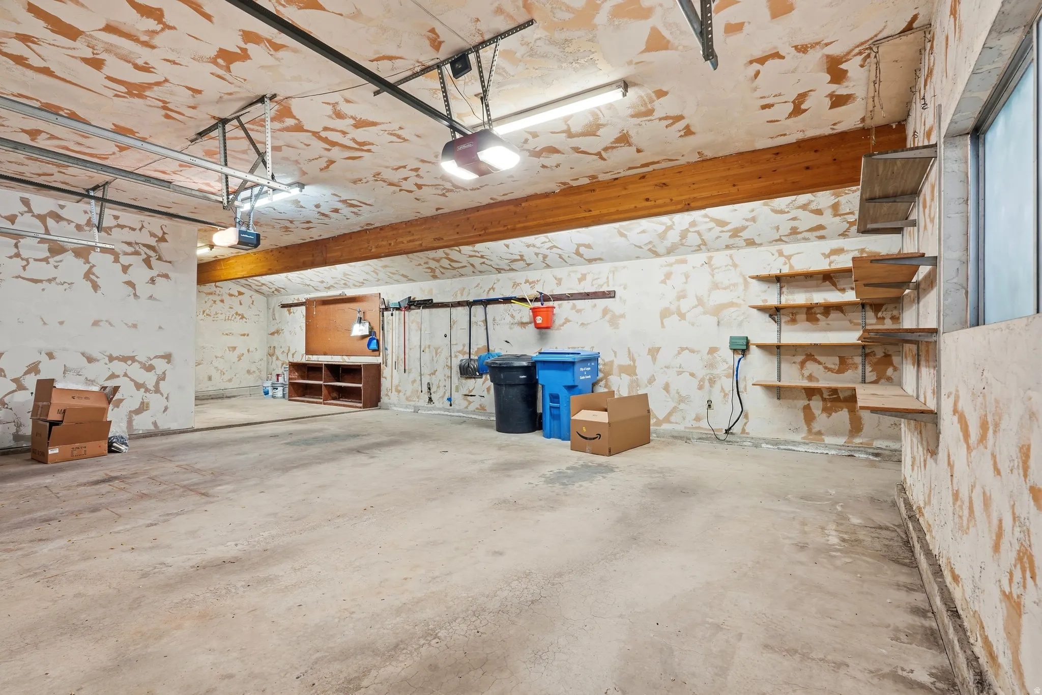 Spacious garage featuring concrete flooring, an exposed wood beam, built-in shelving, and a wall-mounted workbench