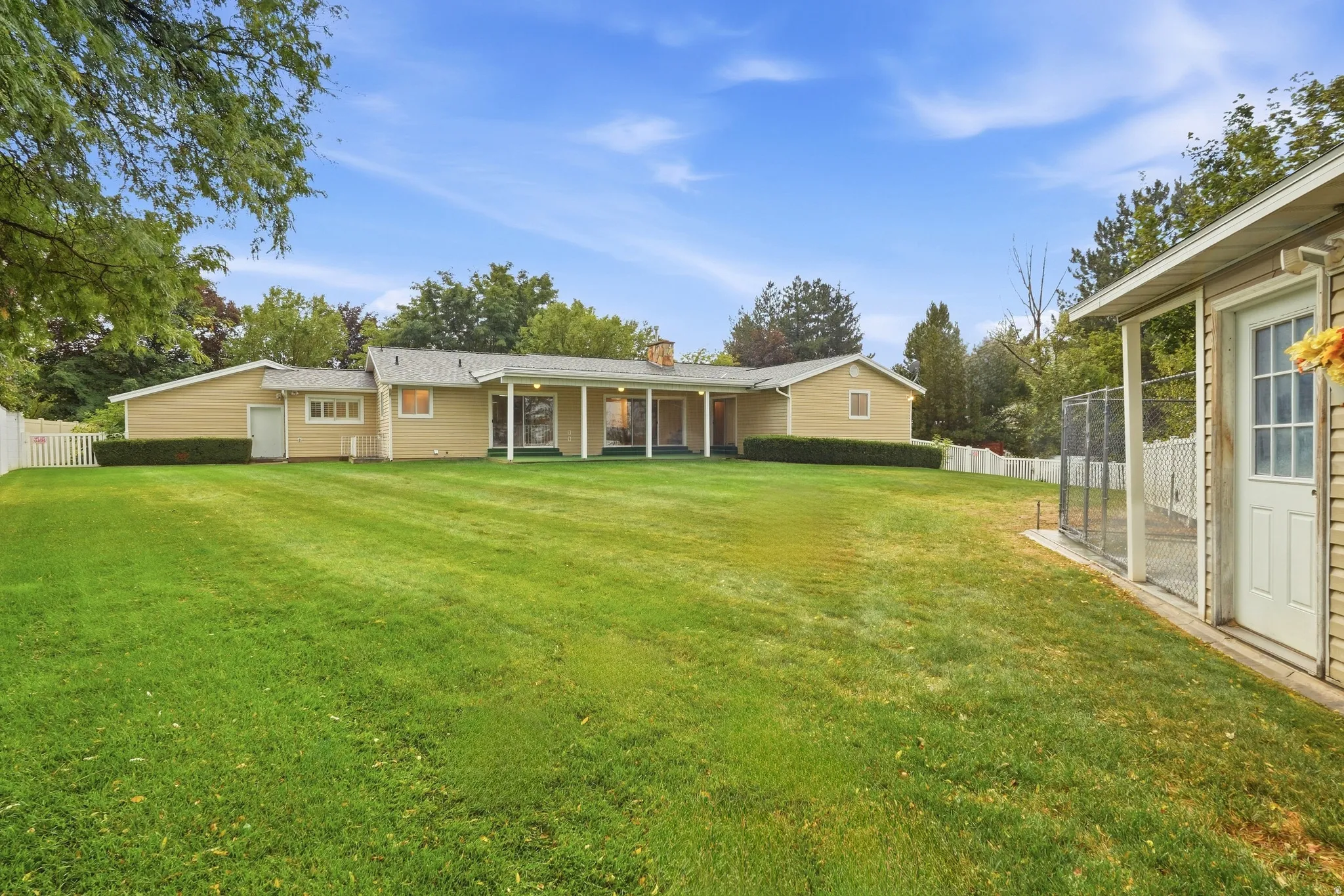 Expansive lawn with a white picket fence, mature trees, a covered patio, and a separate outbuilding