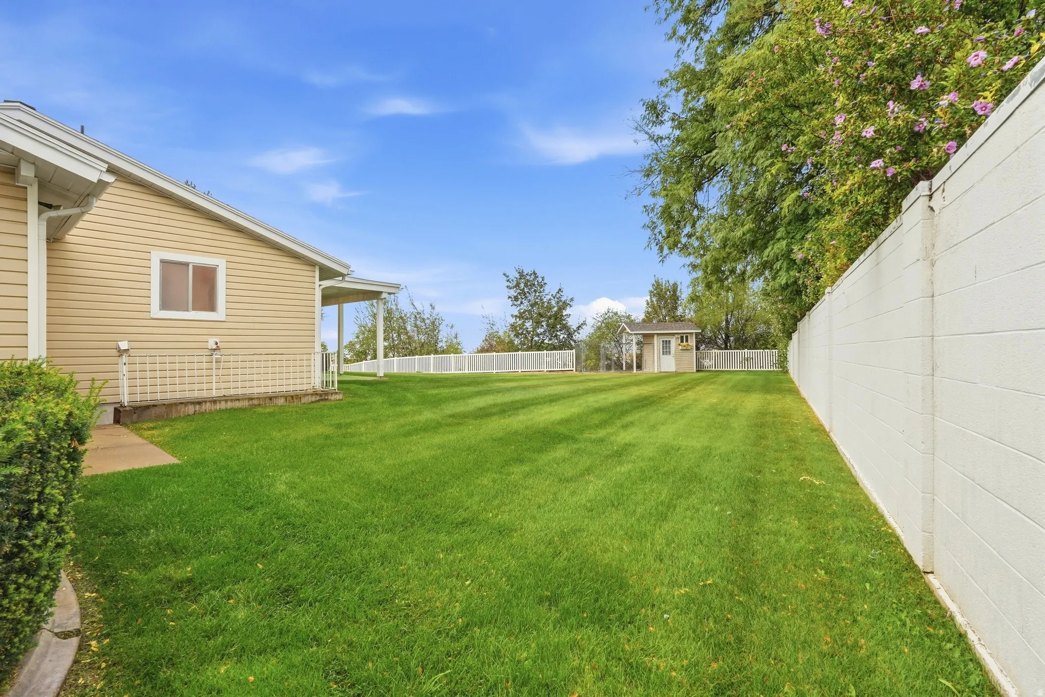 Expansive backyard featuring a meticulously manicured lawn, mature shade trees, and white vinyl fencing