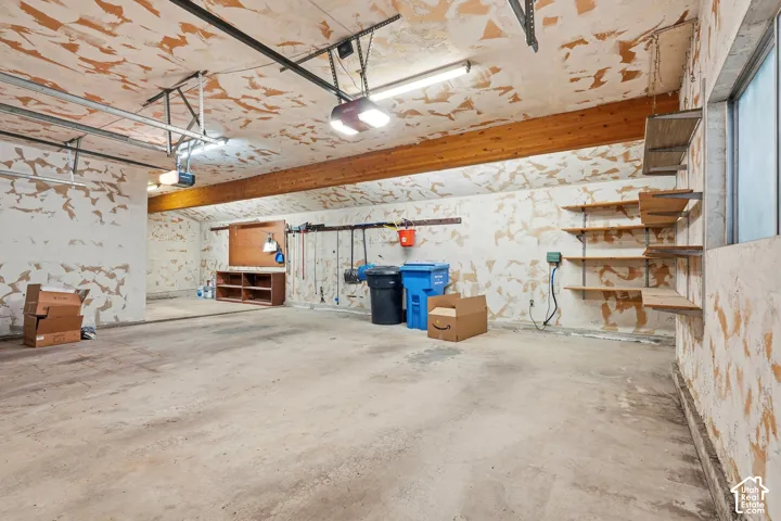 Spacious garage featuring concrete flooring, an exposed wood beam, built-in shelving, and a wall-mounted workbench