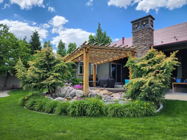 Expansive backyard featuring a manicured lawn, established landscaping, a custom wood pergola, and a stone-clad chimney