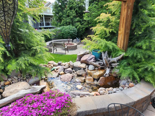 Landscaped outdoor area featuring a custom water feature, stone retaining walls, lush plantings, and a built-in fire pit