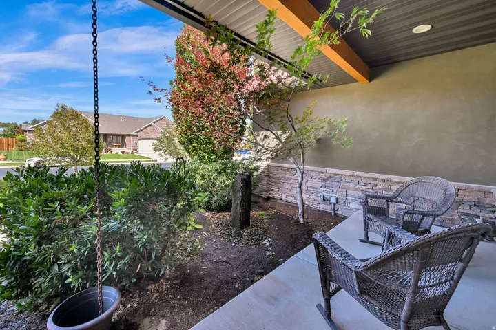 Covered patio with exposed beam ceiling and recessed lighting overlooking a landscaped backyard featuring mature shrubs, ornamental plantings, and woven outdoor seating areas