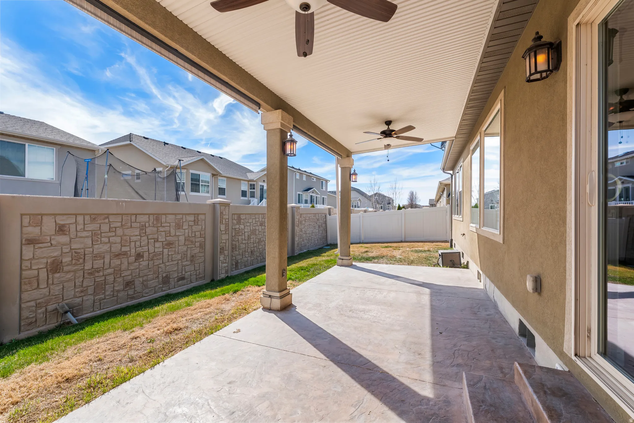 Covered concrete patio with two ceiling fans, exterior sconce lighting, and a textured privacy fence