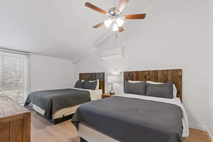 Vaulted ceiling, wood-finish flooring, ceiling fan with integrated lighting, wall-mounted air conditioning unit, and wood-finish headboards