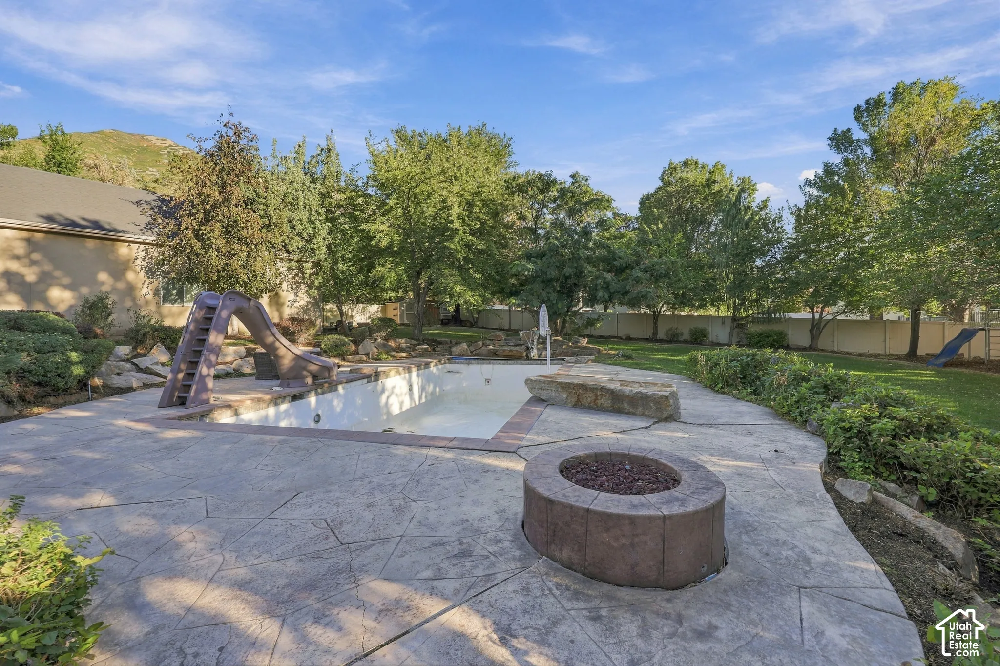 Stamped concrete patio with integrated fire pit, expansive lawn, mature trees, and an in-ground swimming pool featuring a slide