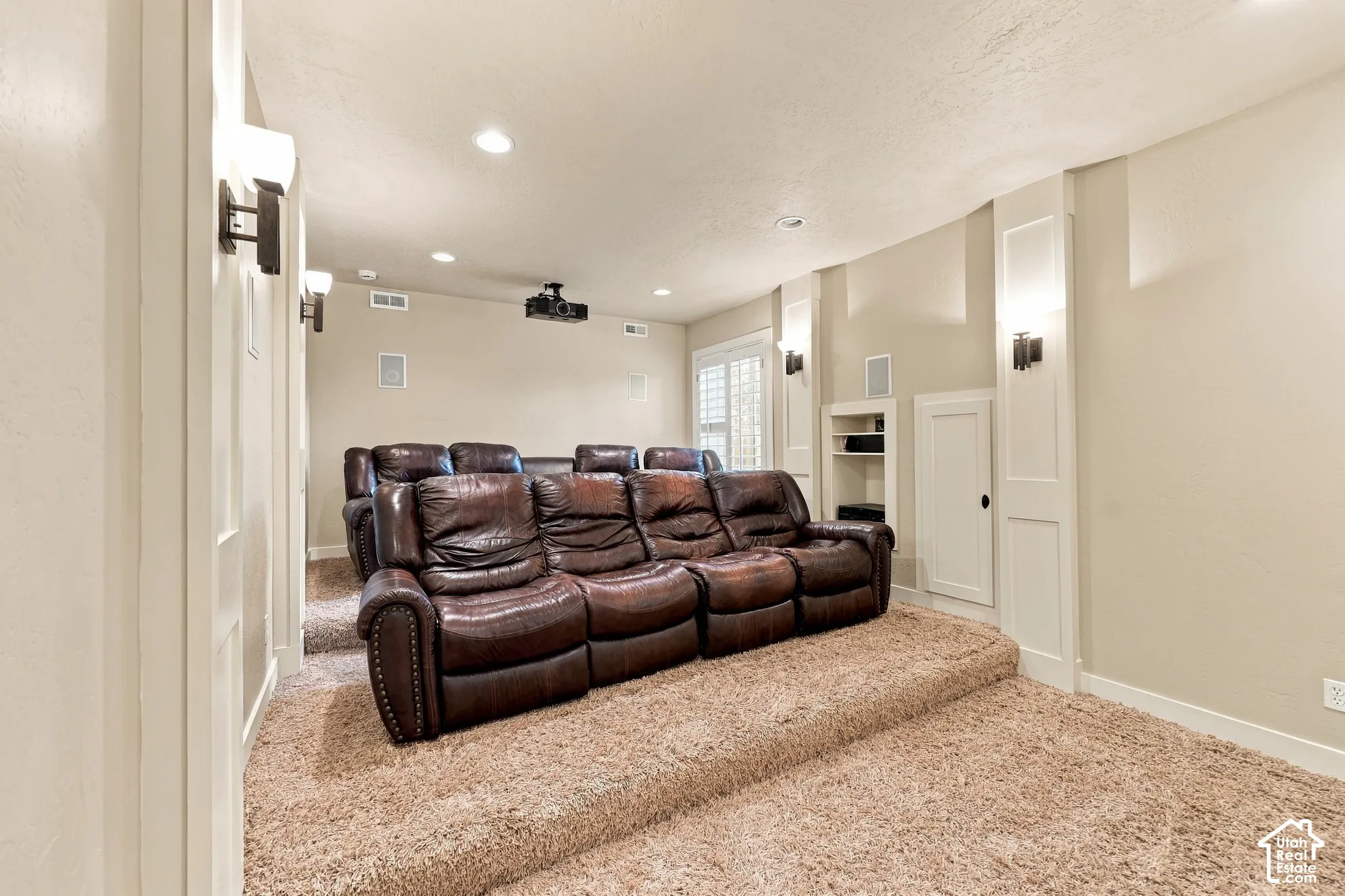 Tiered flooring with plush carpeting, recessed ceiling lighting, wall-mounted sconces, built-in shelving, and a ceiling-mounted projector