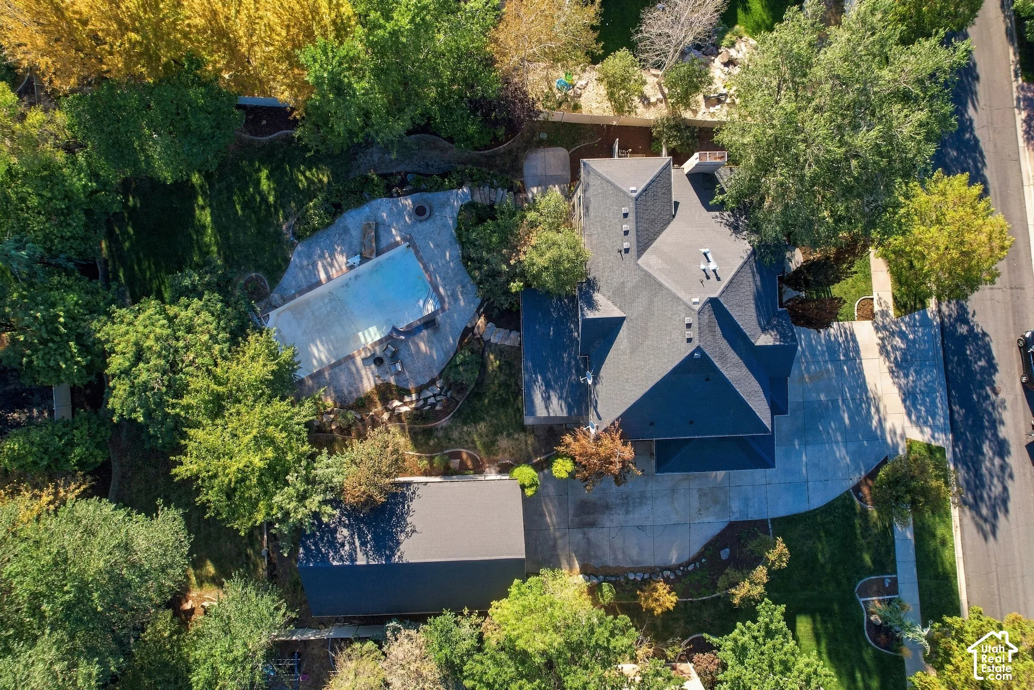 Pitched rooflines with dark shingles, expansive paved driveway, detached garage, in-ground swimming pool with surrounding hardscaping, and mature landscaping