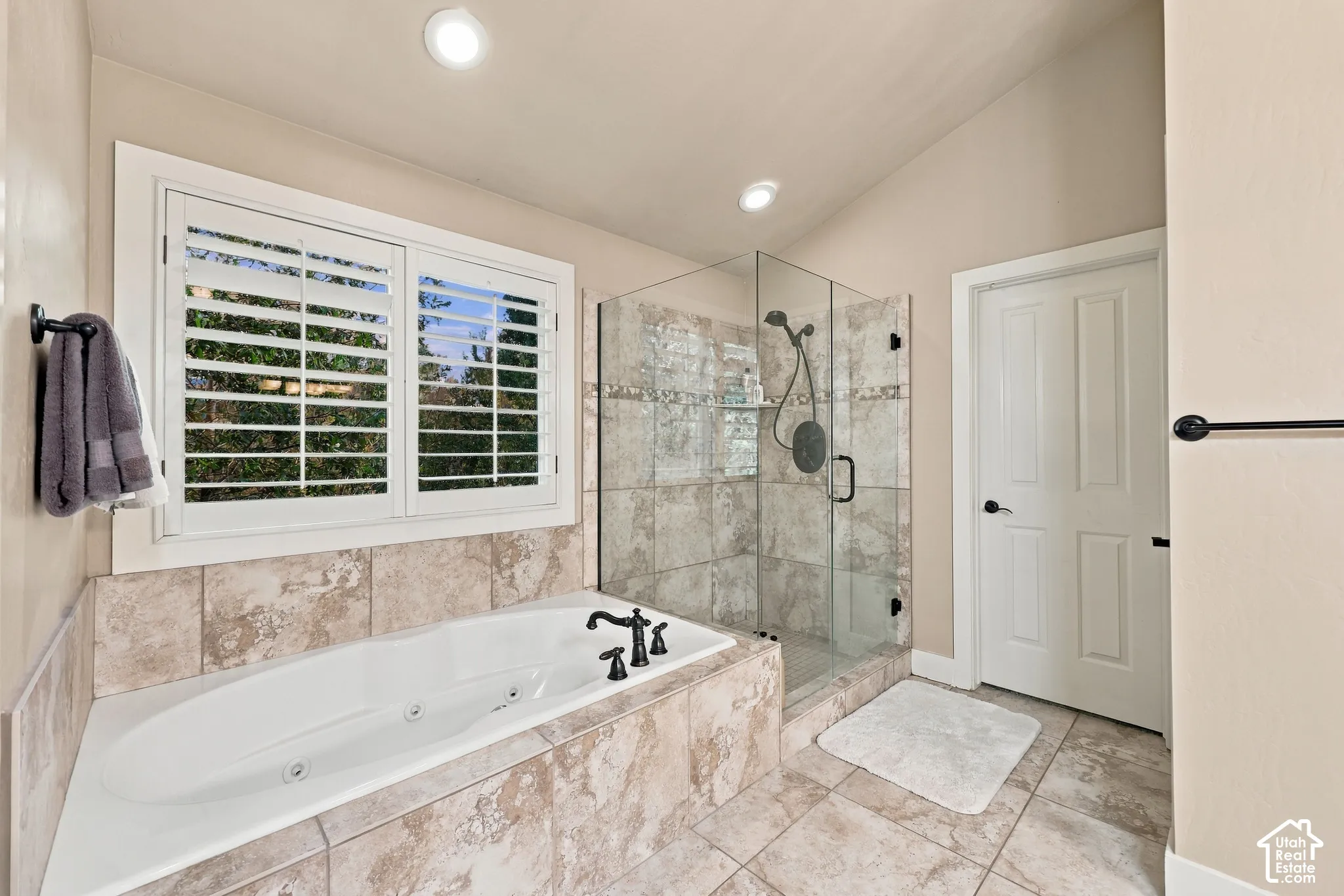 Recessed lighting illuminates a built-in jetted tub with tile surround, a glass-enclosed shower with tile walls, and a window featuring white plantation shutters