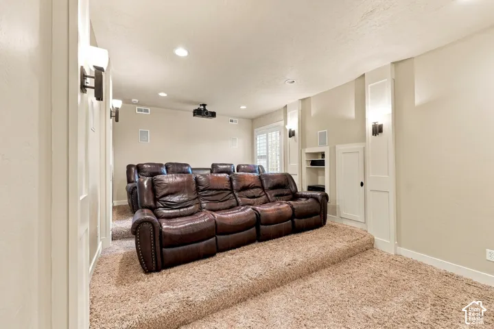 Tiered flooring with plush carpeting, recessed ceiling lighting, wall-mounted sconces, built-in shelving, and a ceiling-mounted projector