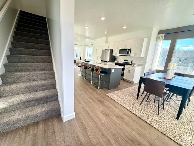Open-concept living area featuring wood-finish flooring, a kitchen island with seating, white cabinetry, stainless steel appliances, and a dining area with expansive windows