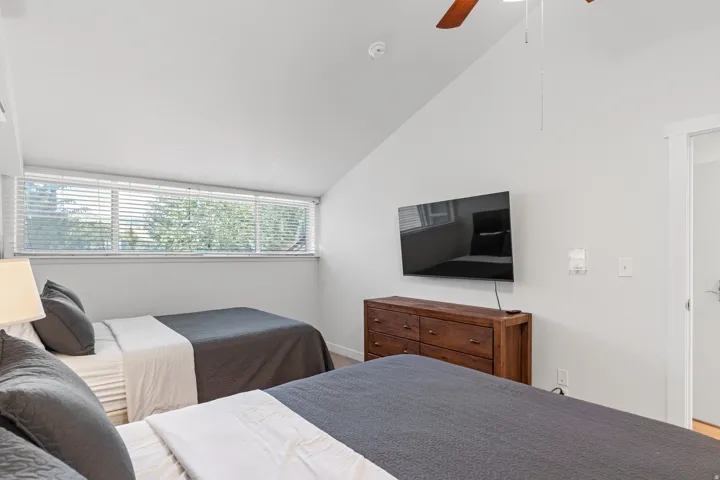 Sloped ceiling room featuring a long horizontal window with blinds, wood-finish flooring, a ceiling fan, and a wall-mounted flat-screen display